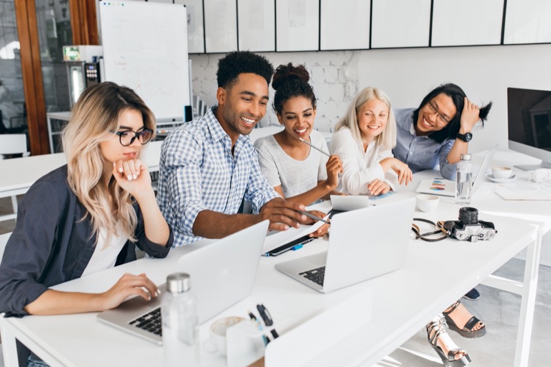 smiling african student pointing with pencil at laptop screen concentrated blonde woman in glasses propping chin with hand while working with computer in office